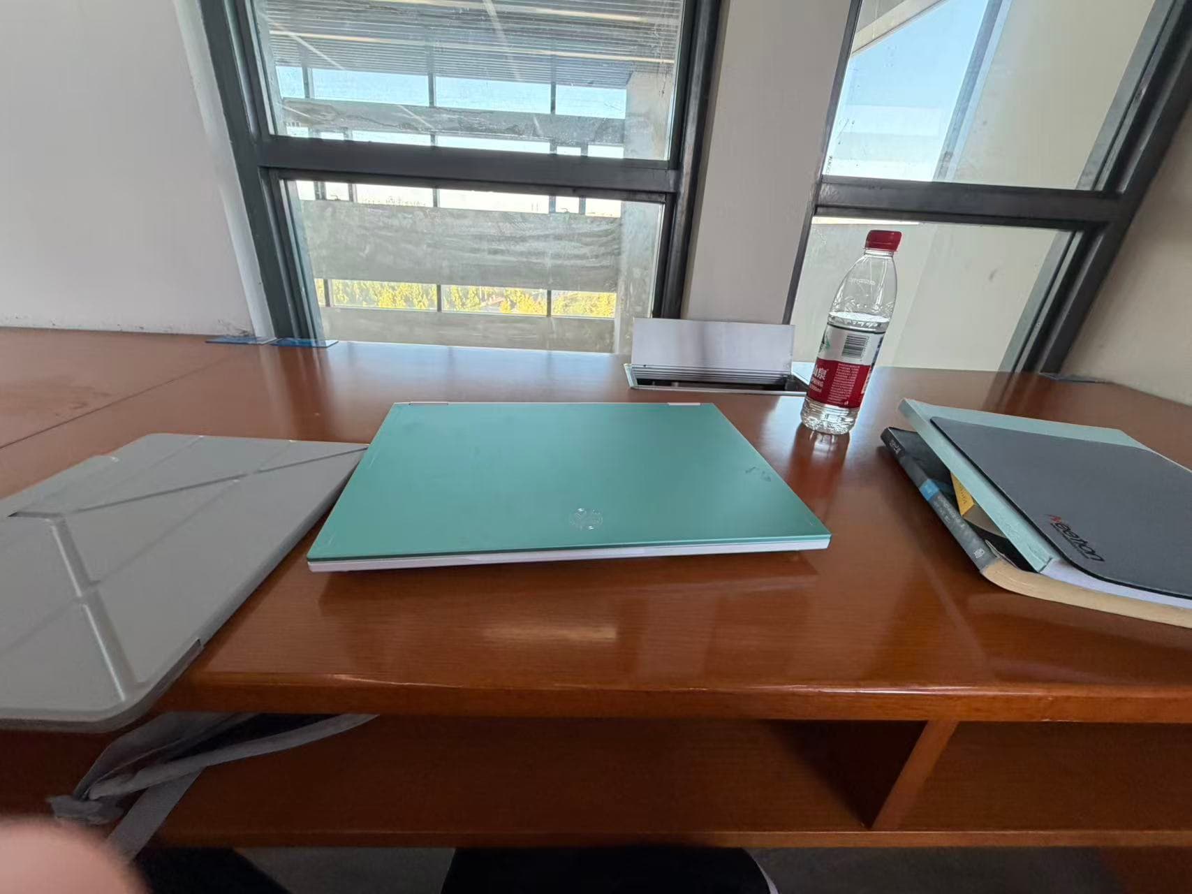 A laptop, books, and a tablet on a wooden table beside a library window