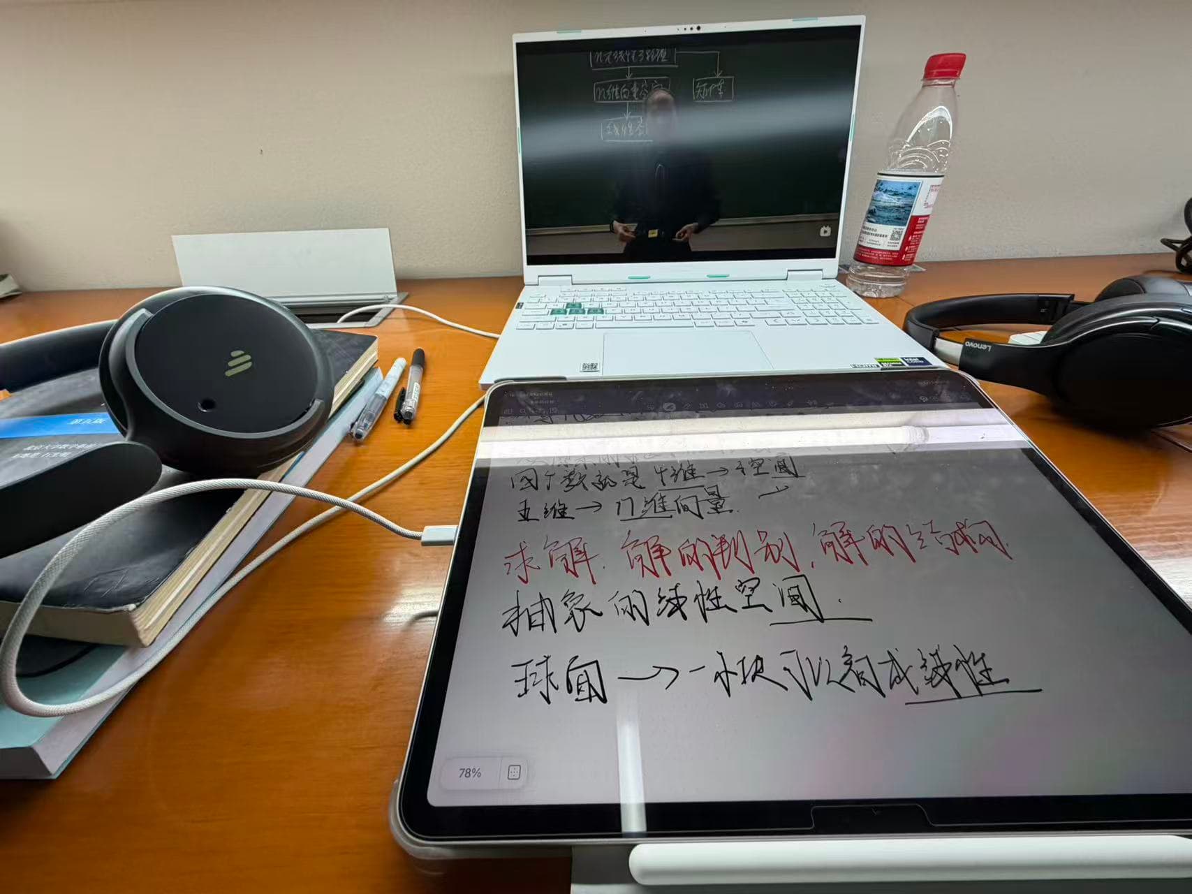 A laptop, tablet, notes, and headphones on a library desk, with a linear algebra lecture playing on the laptop