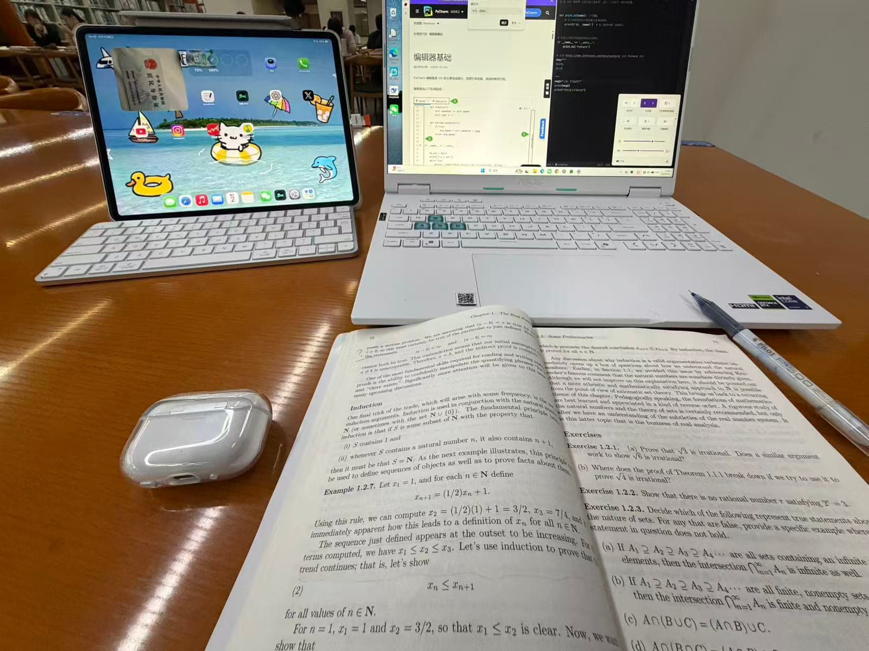 An open mathematics textbook, laptop, tablet, and earphones on a library desk
