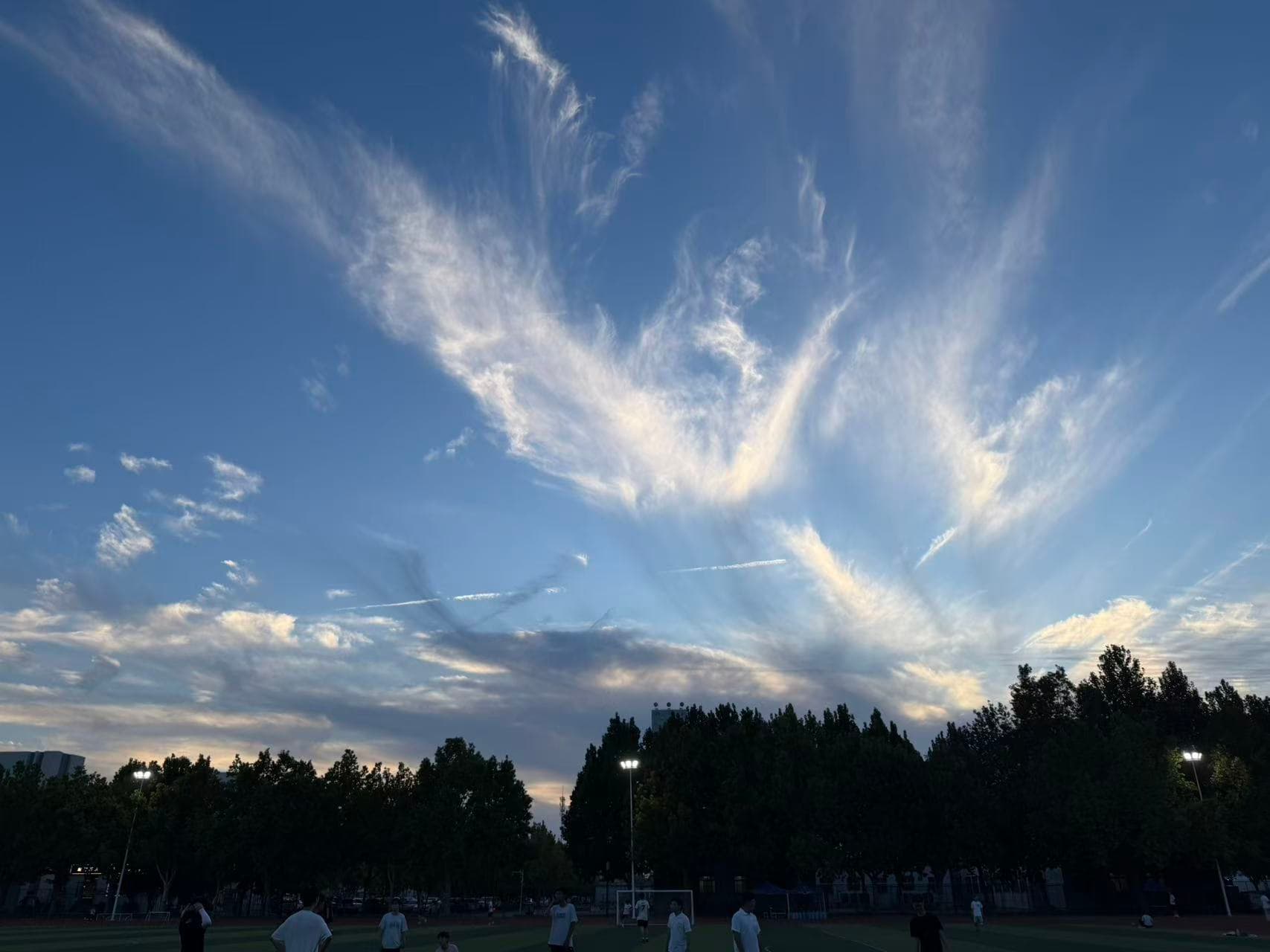 White and pale gold evening clouds spread above the football field, with the floodlights already on beneath the trees