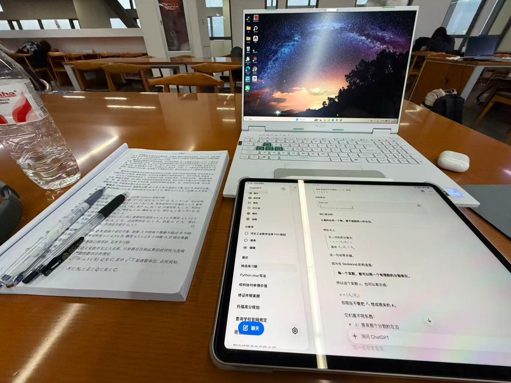A mathematical analysis textbook, laptop, and tablet spread across a wooden library table
