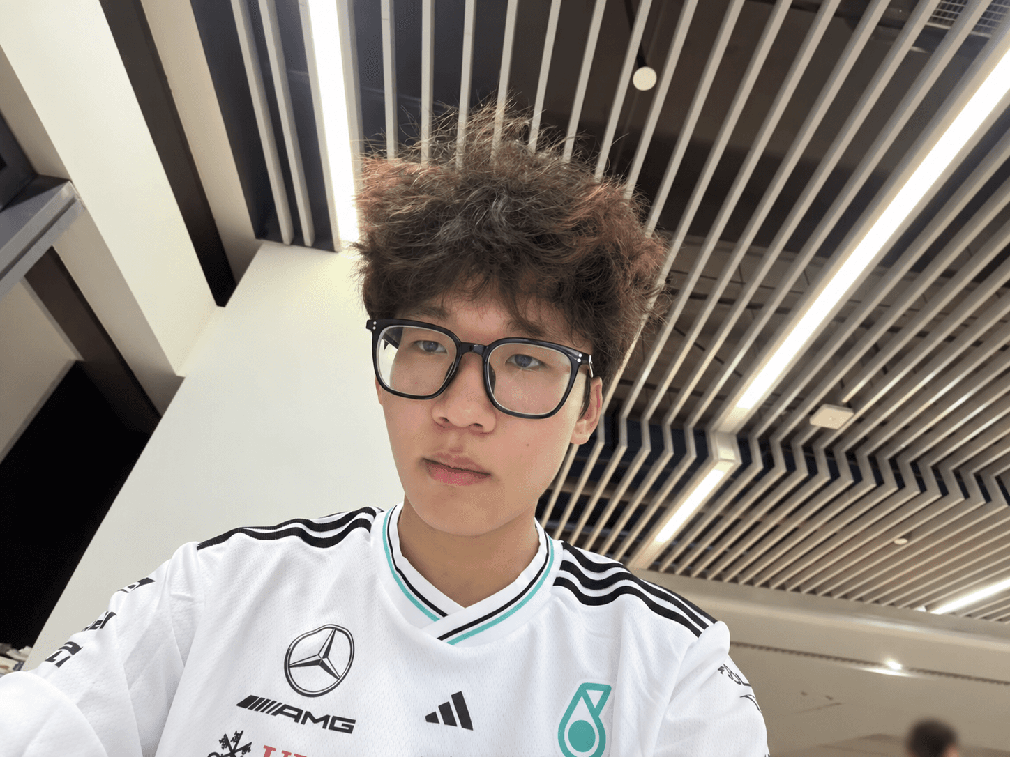 A selfie in the library under ceiling lights, wearing black-framed glasses and a white sports shirt