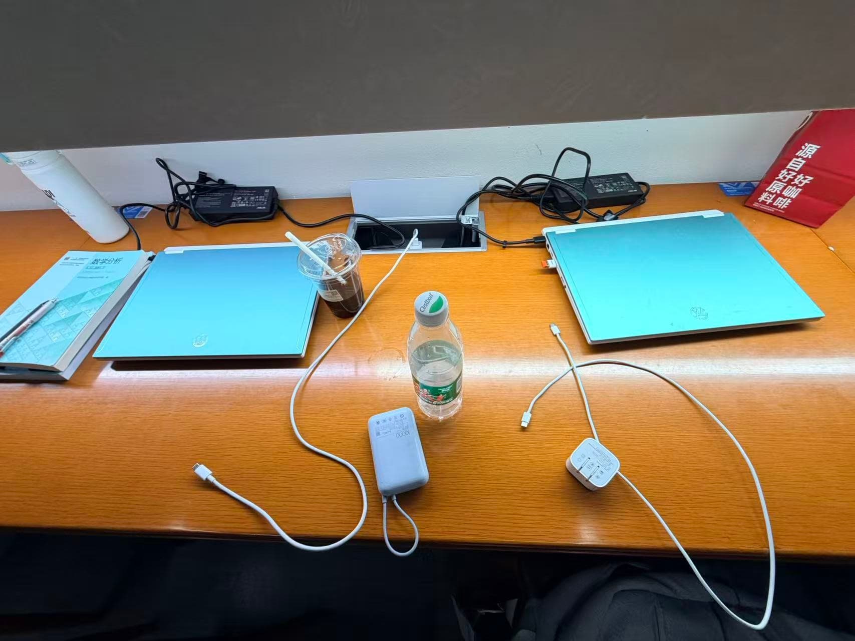 Laptops, books, water, and charging cables spread across a long library desk