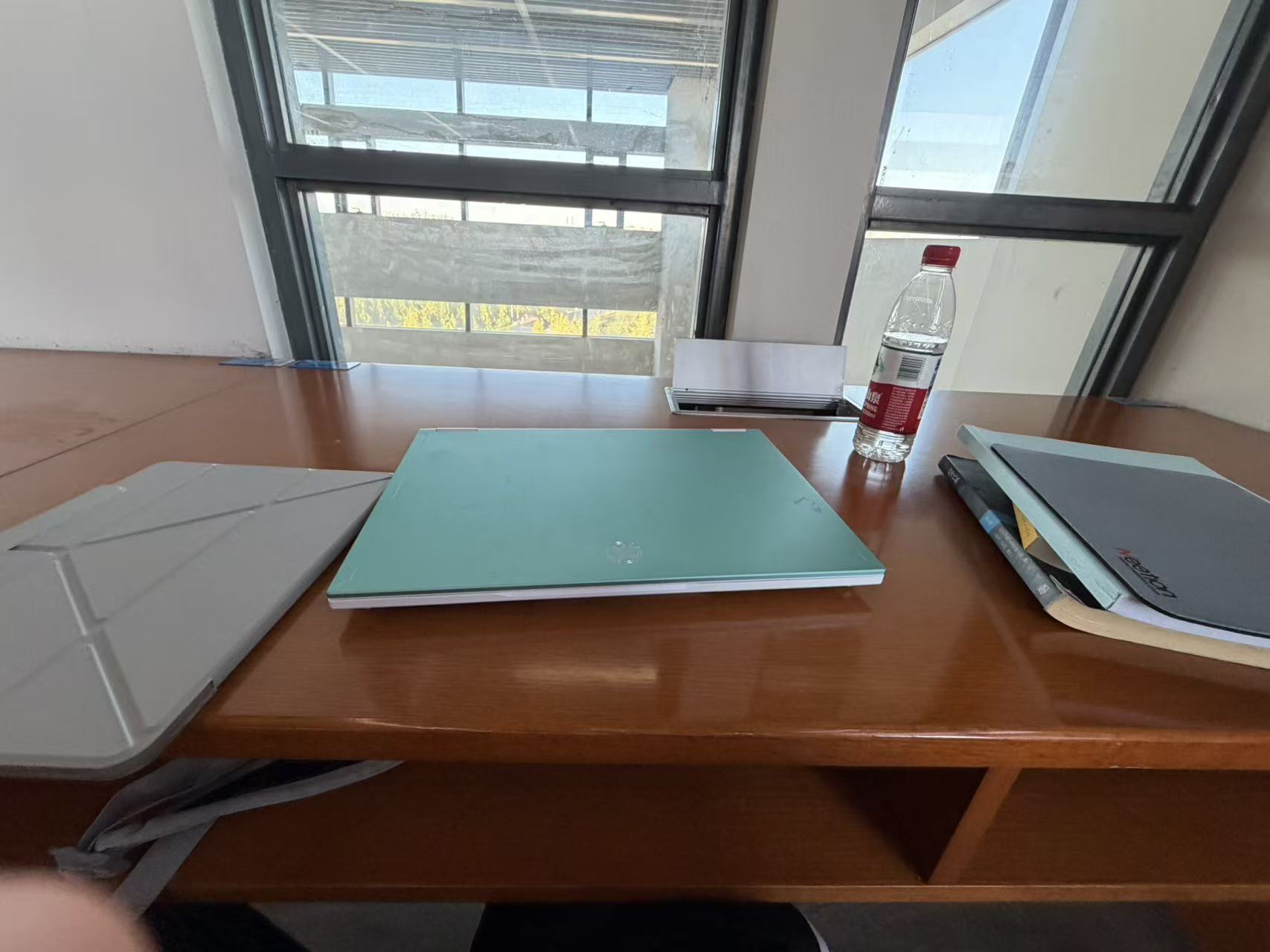 A laptop, books, and a tablet on a wooden table beside a library window