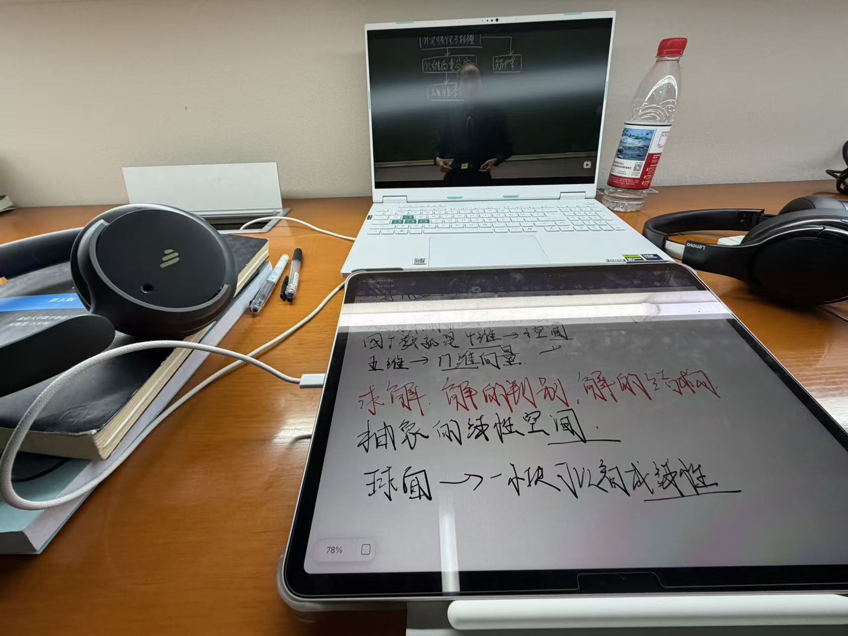 A laptop, tablet, notes, and headphones on a library desk, with a linear algebra lecture playing on the laptop
