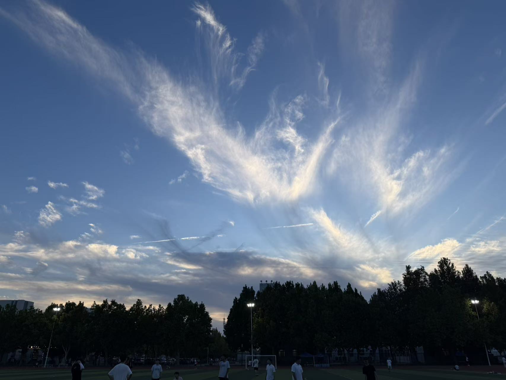 White and pale gold evening clouds spread above the football field, with the floodlights already on beneath the trees