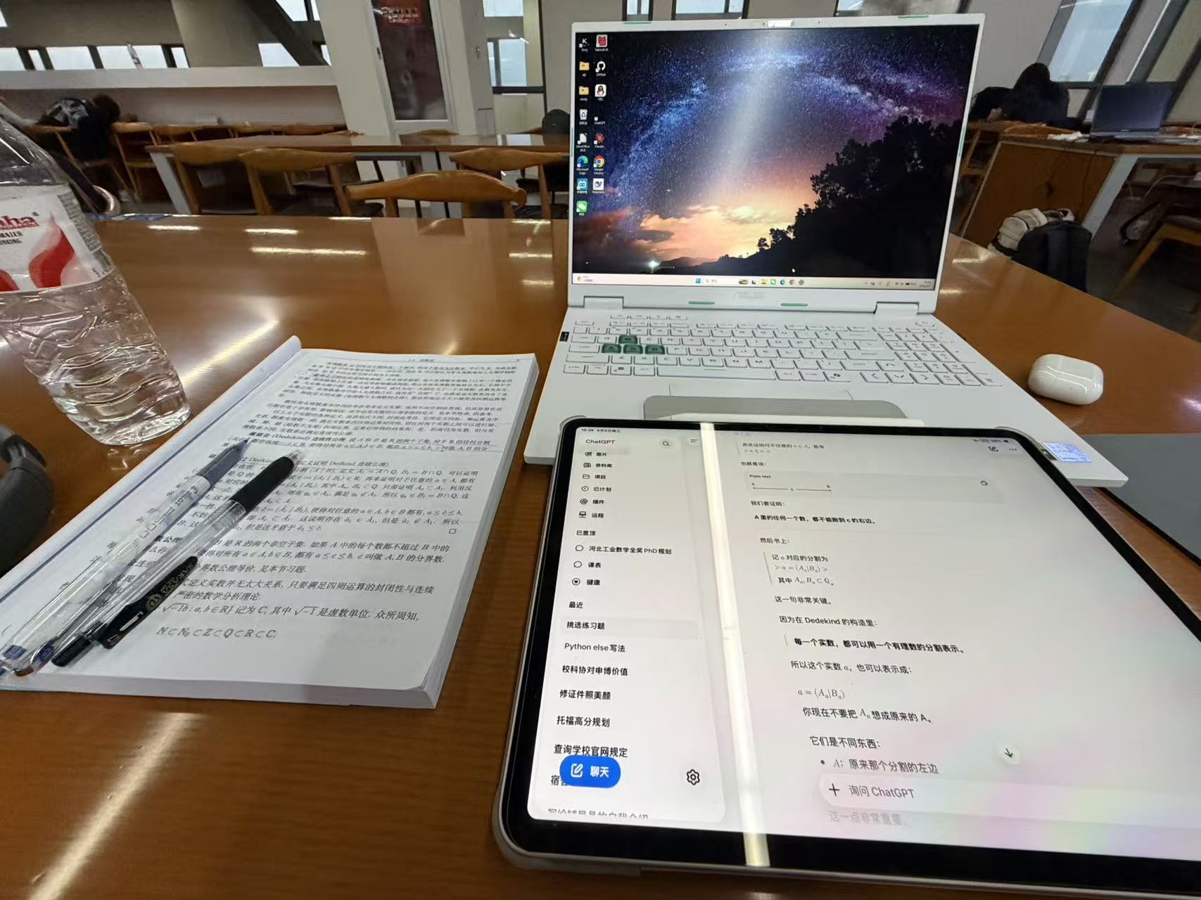 A mathematical analysis textbook, laptop, and tablet spread across a wooden library table